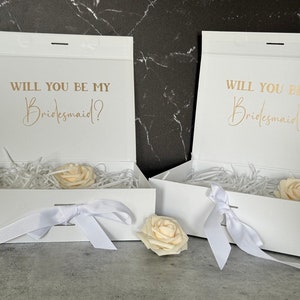 May include: Two white gift boxes with white ribbon bows and a single white rose on top of each box. The boxes are open and have white tissue paper inside. The lids of the boxes have the text "Will you be my Bridesmaid?" in gold lettering.