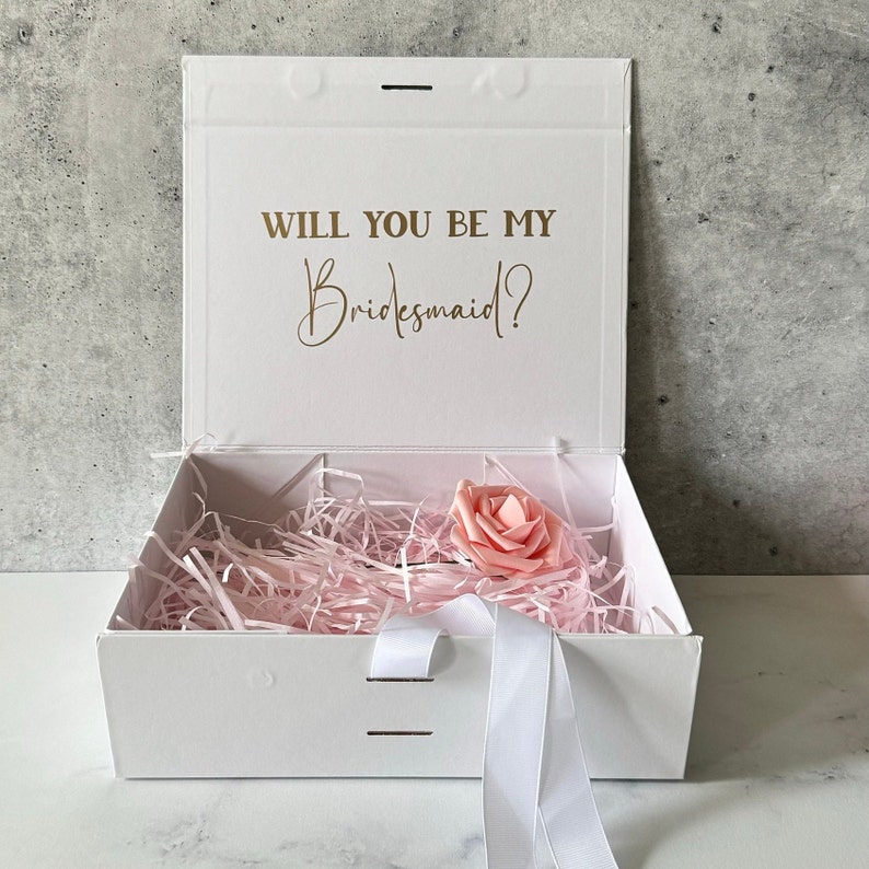 May include: White gift box with a pink rose inside. The lid of the box has the text "Will you be my Bridesmaid?" in gold lettering.