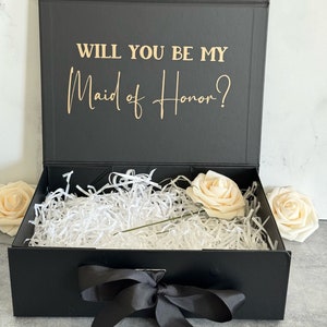 May include: A black rectangular gift box with a hinged lid featuring the gold text "WILL YOU BE MY Maid of Honor?" inside. The box is filled with white shredded paper filler and contains a single cream-colored artificial rose with a green stem.