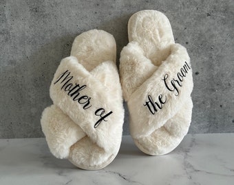 Peut inclure: Une paire de chaussons en fausse fourrure crème avec une bride croisée. Le chausson gauche porte le texte en écriture noire Mother of et le chausson droit porte le texte en écriture noire the Groom, imprimés sur le tissu en peluche.