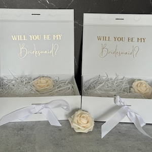 May include: Two white gift boxes with white ribbon bows and a single white rose on top of each box. The boxes have the text "Will you be my Bridesmaid?" printed on the top in gold lettering.