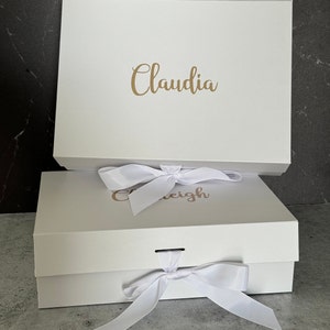 Peut inclure: Deux bo&icirc;tes cadeaux blanches avec des n&oelig;uds en ruban blanc. La bo&icirc;te du haut porte le nom "Claudia" &eacute;crit en lettres dor&eacute;es. La bo&icirc;te du bas porte le nom "Caleigh" &eacute;crit en lettres dor&eacute;es.
