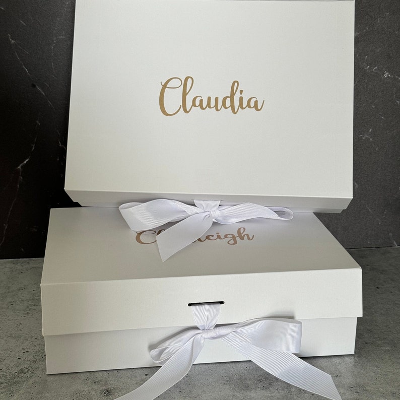 Peut inclure: Deux bo&icirc;tes cadeaux blanches avec des n&oelig;uds en ruban blanc. La bo&icirc;te du haut porte le nom "Claudia" &eacute;crit en lettres dor&eacute;es. La bo&icirc;te du bas porte le nom "Caleigh" &eacute;crit en lettres dor&eacute;es.
