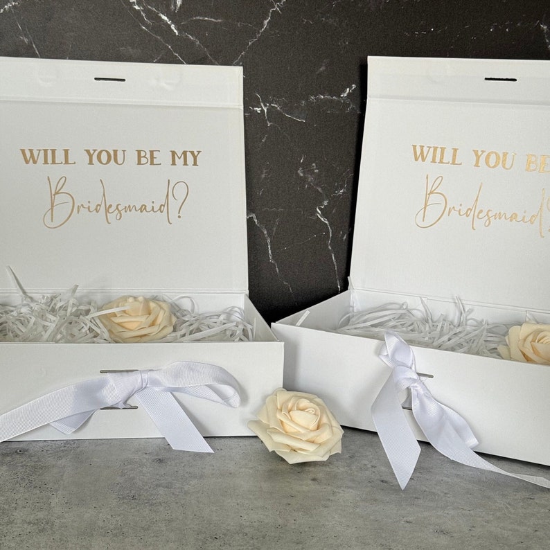 May include: Two white gift boxes with white ribbon bows and a single white rose on top of each box. The boxes are open and have white tissue paper inside. The lids of the boxes have the text "Will you be my Bridesmaid?" in gold lettering.