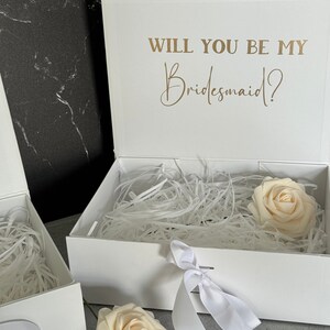 Peut inclure: Bo&icirc;te cadeau blanche avec un ruban de satin blanc et une seule rose blanche &agrave; l'int&eacute;rieur. Le couvercle de la bo&icirc;te porte le texte "Veux-tu &ecirc;tre ma demoiselle d'honneur ?" en lettres dor&eacute;es.