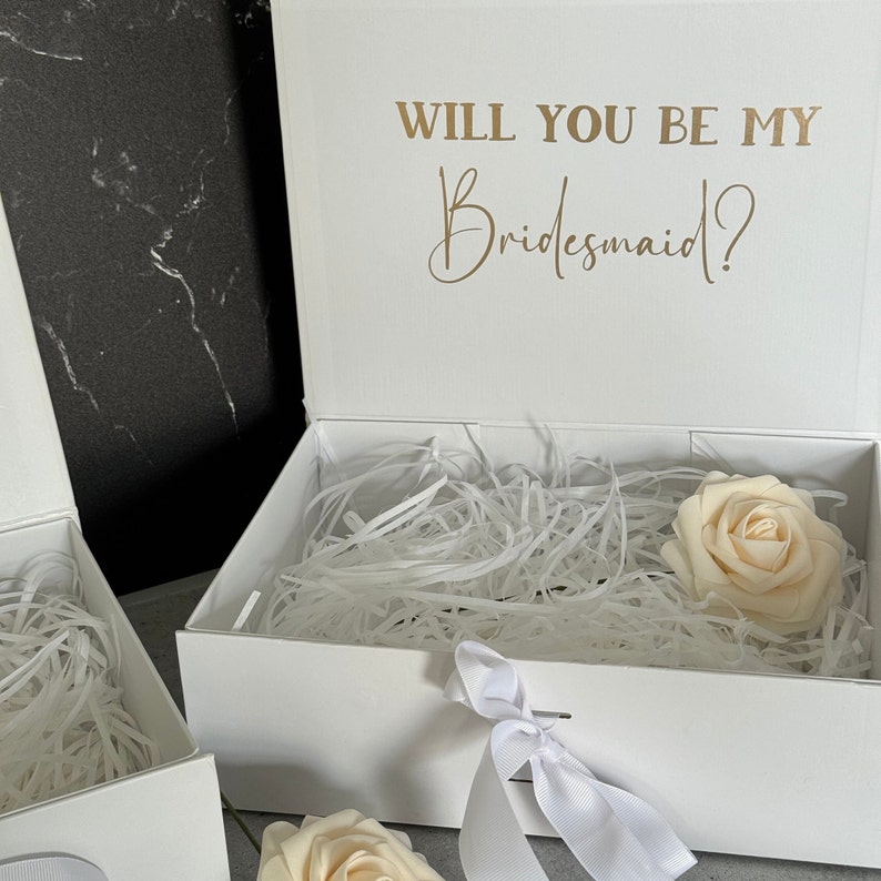 Peut inclure: Bo&icirc;te cadeau blanche avec un ruban de satin blanc et une seule rose blanche &agrave; l'int&eacute;rieur. Le couvercle de la bo&icirc;te porte le texte "Veux-tu &ecirc;tre ma demoiselle d'honneur ?" en lettres dor&eacute;es.