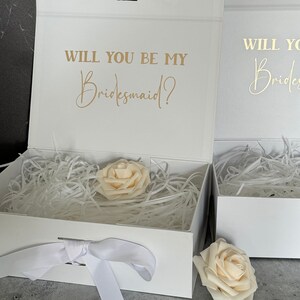 Peut inclure: Bo&icirc;te cadeau blanche avec un ruban de satin blanc et une seule rose blanche. La bo&icirc;te est ouverte et l'int&eacute;rieur est doubl&eacute; de papier de soie blanc. Le couvercle de la bo&icirc;te porte le texte "Veux-tu &ecirc;tre ma demoiselle d'honneur ?" en lettres dor&eacute;es.