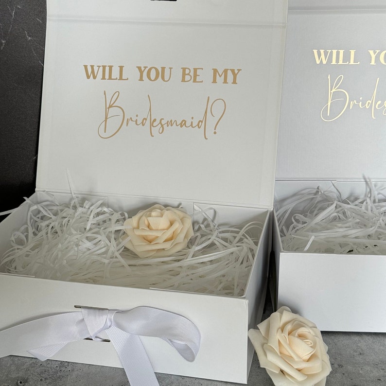 Peut inclure: Bo&icirc;te cadeau blanche avec un ruban de satin blanc et une seule rose blanche. La bo&icirc;te est ouverte et l'int&eacute;rieur est doubl&eacute; de papier de soie blanc. Le couvercle de la bo&icirc;te porte le texte "Veux-tu &ecirc;tre ma demoiselle d'honneur ?" en lettres dor&eacute;es.