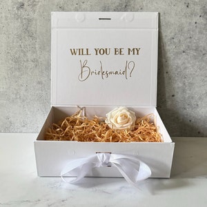 Peut inclure: Bo&icirc;te cadeau blanche avec un ruban de satin blanc et une seule rose blanche. Le couvercle de la bo&icirc;te porte le texte "Veux-tu &ecirc;tre ma demoiselle d'honneur ?" en lettres dor&eacute;es.
