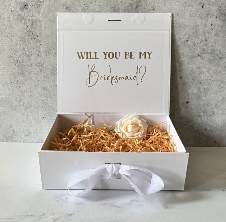 Peut inclure: Bo&icirc;te cadeau blanche avec un ruban de satin blanc et une seule rose blanche. Le couvercle de la bo&icirc;te porte le texte "Veux-tu &ecirc;tre ma demoiselle d'honneur ?" en lettres dor&eacute;es.
