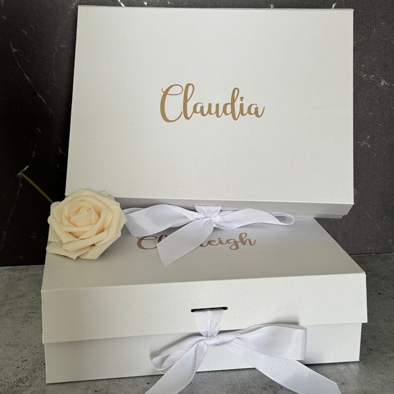 Peut inclure: Deux bo&icirc;tes cadeaux blanches avec des lettres dor&eacute;es. La bo&icirc;te du haut dit "Claudia" et la bo&icirc;te du bas dit "Caleigh". Les deux bo&icirc;tes ont des n&oelig;uds en ruban de satin blanc.