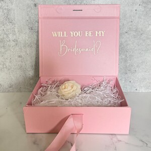 Peut inclure: Bo&icirc;te cadeau rose avec un ruban de satin blanc et une seule rose blanche &agrave; l'int&eacute;rieur. Le couvercle de la bo&icirc;te porte le texte "Veux-tu &ecirc;tre ma demoiselle d'honneur ?" en lettres blanches.