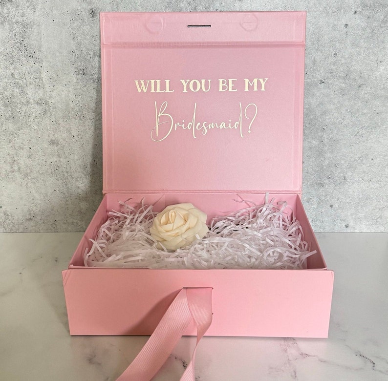 Peut inclure: Bo&icirc;te cadeau rose avec un ruban de satin blanc et une seule rose blanche &agrave; l'int&eacute;rieur. Le couvercle de la bo&icirc;te porte le texte "Veux-tu &ecirc;tre ma demoiselle d'honneur ?" en lettres blanches.