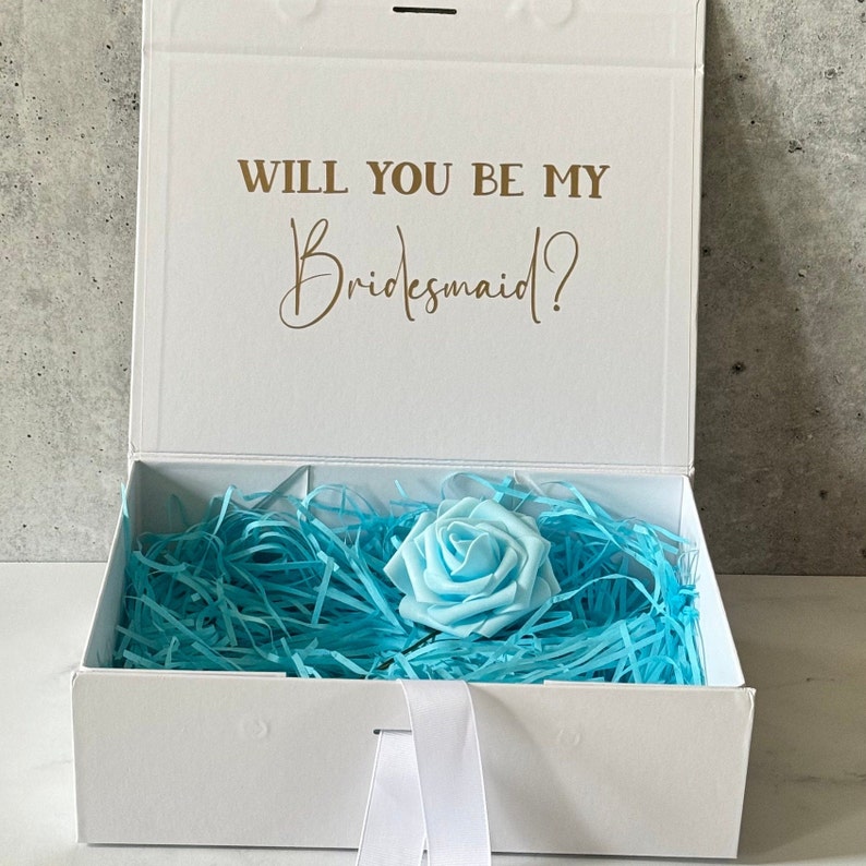 Peut inclure: Bo&icirc;te blanche avec un ruban bleu et une rose bleu clair &agrave; l'int&eacute;rieur. La bo&icirc;te porte l'inscription "Veux-tu &ecirc;tre ma demoiselle d'honneur ?" sur le couvercle.