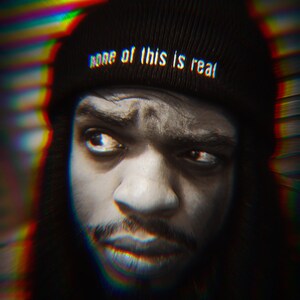Puede incluir: Primer plano de una persona que lleva una boina negra con el texto "None of this is real" en blanco. La persona tiene barba y mira hacia abajo.