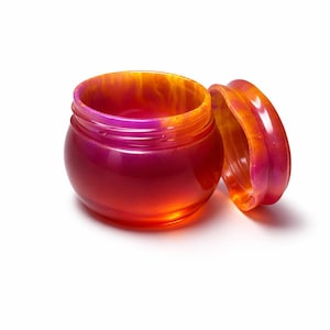May include: A small, round jar with a matching lid, both in a gradient of colors. The jar transitions from a deep red at the base to a vibrant orange and pink at the rim. The lid is placed next to the jar, showing the same color scheme.