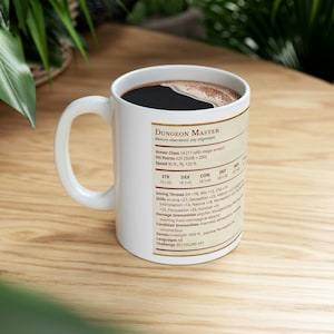 Puede incluir: Taza de cerámica blanca con café negro en su interior. La taza tiene un borde negro y un gráfico blanco con el texto "Dungeon Master" y un bloque de estadísticas de personaje de Dungeons and Dragons.