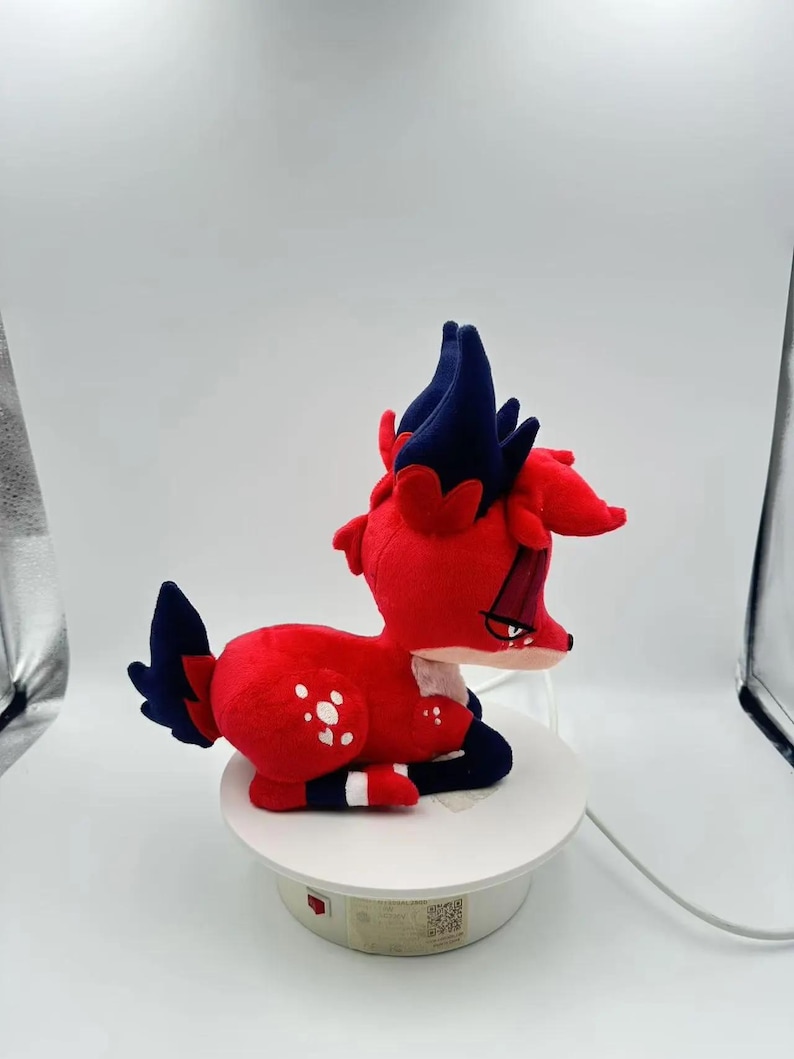 Puede incluir: Un peluche con forma de criatura roja y azul parecida a un ciervo. El juguete tiene un cuerpo rojo con manchas blancas, detalles azules y un dise&ntilde;o estilizado. Est&aacute; tumbado sobre una superficie blanca.