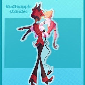 May include: A digital illustration of a Radioapple standee featuring two animated characters. One character with red hair and a red vest carries another character with blonde hair and a pink vest. The text KanmuruDesigns and Radioapple standee is visible.