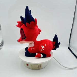 Puede incluir: Un peluche de ciervo rojo con detalles en azul oscuro en las orejas y la cola. El ciervo tiene manchas blancas y un parche blanco en el pecho. Est&aacute; posado sobre una plataforma circular blanca. El juguete es de tela suave.
