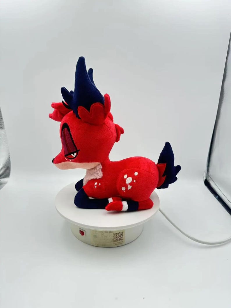 Puede incluir: Un peluche de ciervo rojo con detalles en azul oscuro en las orejas y la cola. El ciervo tiene manchas blancas y un parche blanco en el pecho. Est&aacute; posado sobre una plataforma circular blanca. El juguete es de tela suave.