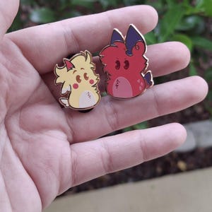 May include: Two enamel pins featuring cartoon-style creatures. One is yellow with a white belly, the other is red with purple accents. Both pins have black pin backs and are held in a hand.