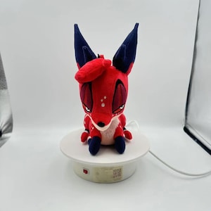 Puede incluir: Un peluche de zorro rojo con orejas y patas azul oscuro y lunares blancos. El zorro tiene ojos grandes y est&aacute; colocado sobre una plataforma giratoria blanca. El juguete tiene un dise&ntilde;o de dibujos animados.