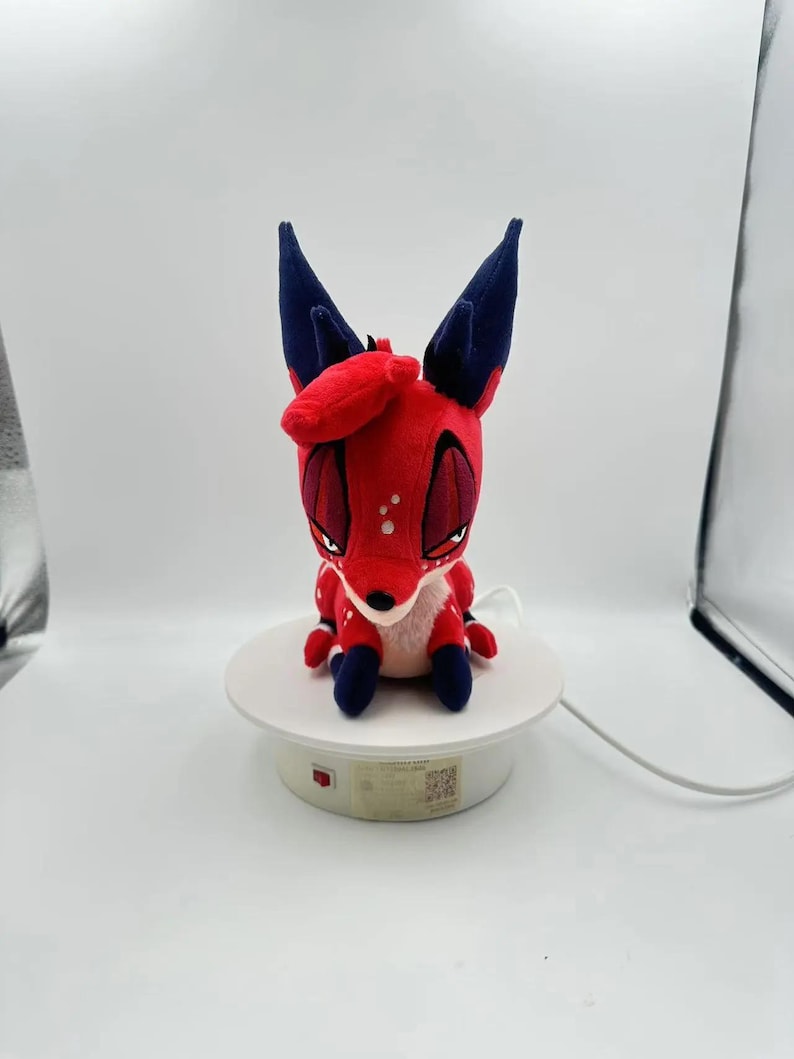 Puede incluir: Un peluche de zorro rojo con orejas y patas azul oscuro y lunares blancos. El zorro tiene ojos grandes y est&aacute; colocado sobre una plataforma giratoria blanca. El juguete tiene un dise&ntilde;o de dibujos animados.