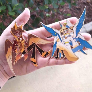 Puede incluir: Dos pines esmaltados con personajes de dibujos animados con alas. Uno tiene un esquema de color marrón y naranja, el otro azul y blanco. Ambos pines tienen contornos dorados.