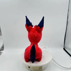 Puede incluir: Un peluche rojo con forma de criatura, visto desde atr&aacute;s. El juguete tiene detalles en azul oscuro en las orejas y la cola. Est&aacute; colocado sobre una plataforma giratoria blanca, sobre un fondo blanco.