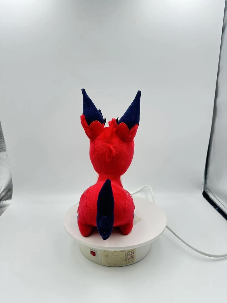 Puede incluir: Un peluche rojo con forma de criatura, visto desde atr&aacute;s. El juguete tiene detalles en azul oscuro en las orejas y la cola. Est&aacute; colocado sobre una plataforma giratoria blanca, sobre un fondo blanco.