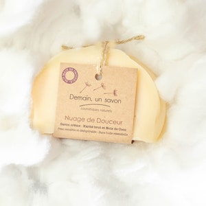 Puede incluir: Una barra de jabón redonda de color amarillo claro envuelta en papel marrón con una etiqueta que dice "Demain, un savon" y "Nuage de Douceur". La etiqueta también dice "Savon crème - Karité brut et Noix de Coco" y "Peau sensible et déshydratée - Sans huile essentielle".