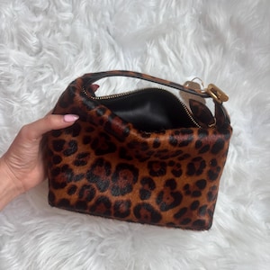 May include: A small handbag featuring a brown and black leopard print pattern with a textured hair-on-hide finish. The bag has a single top handle with a gold-toned metal buckle and a black interior lining with a top zipper closure.