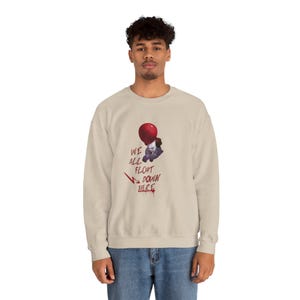 May include: Beige sweatshirt featuring a graphic of a red balloon and a clown with the text "WE ALL FLOAT DOWN HERE" in black and red. The sweatshirt is a crew neck style.