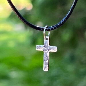 May include: A silver cross pendant on a black cord necklace. The cross has a hammered texture.