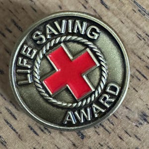 May include: A bronze-colored Life Saving Award pin. The pin features a red cross in the center, surrounded by a rope-like border. The words "LIFE SAVING AWARD" are inscribed around the edge of the pin.