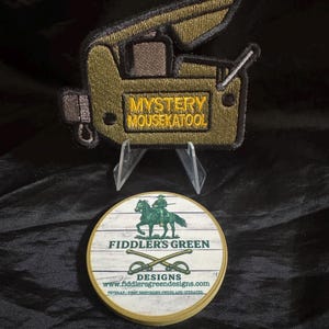 May include: An embroidered patch with the text "MYSTERY MOUSEKATOOL" in yellow, shaped like a vintage camera. Below it, a round coaster with the words "FIDDLERS GREEN DESIGNS" and a cavalry illustration.