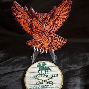 May include: Embroidered patch of an owl with outstretched wings in shades of orange and brown. Below, a round wooden coaster with the text "FIDDLER'S GREEN DESIGNS" and a graphic of a soldier on horseback.
