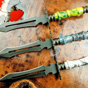 Puede incluir: Tres dagas negras con mangos de diferentes colores. Los mangos están hechos de madera y tienen un patrón de mármol. Las dagas tienen una punta afilada y un borde dentado.