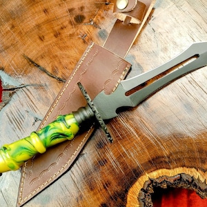 May include: A dagger with a black blade and a handle with yellow and green swirls, resting on a brown leather sheath. The handle has a decorative, ridged design. The sheath is on a wooden surface.