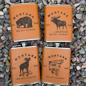 May include: Four rectangular stainless steel flasks wrapped in tan faux leather. Each flask features the text MONTANA, EST 1889, and BIG SKY COUNTRY. The designs include a black engraved illustration of a bear, a moose, a bighorn sheep, and a trout.