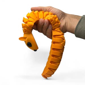 May include: Orange articulated toy shaped like a stylized animal, possibly a squirrel or a similar creature. The toy is held in a hand, showcasing its flexible, segmented design. The toy's head has black eyes and a small, black nose.