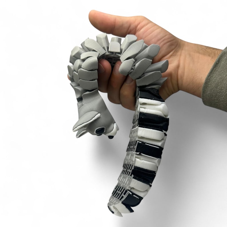 Op de afbeelding: Een grijs gearticuleerd speelgoed in de vorm van een gestileerd dier, vastgehouden in een hand. Het speelgoed heeft een gesegmenteerd lichaam met een kop en een staart. De staart is zwart en wit, terwijl de kop en het lichaam grijs zijn.