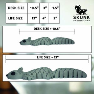 May include: A gray squirrel-shaped desk organizer. The image shows two sizes: desk size at 10.5 inches long and life size at 13 inches long. The organizer has a textured design and is labeled with the text "SKUNK PRINTS."