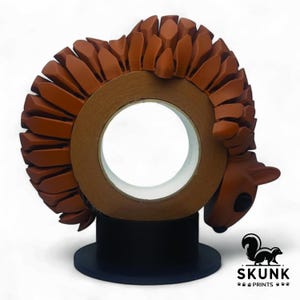 May include: A brown tape dispenser shaped like a squirrel, with a decorative, textured design. The squirrel is perched on a roll of tape, with the tape roll mounted on a black base. The Skunk Prints logo is visible.