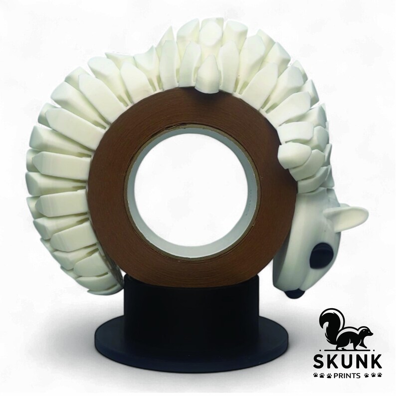 May include: A decorative tape dispenser featuring a white, stylized animal design wrapped around a brown tape roll. The animal has a white body, black eyes, and a small, pointed nose. The dispenser sits on a black base. The Skunk Prints logo is in the bottom right corner.