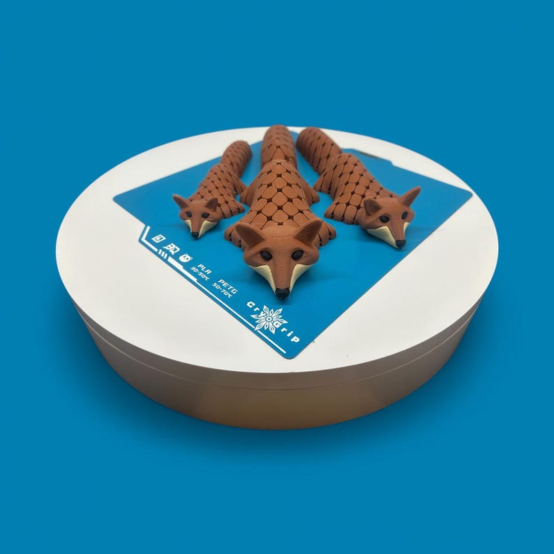 May include: A white, round container with three brown fox figurines on top. The foxes are arranged in a row, with the largest in the center. The container has a blue square with text and a logo. The background is a solid blue.