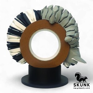Op de afbeelding: Een sculptuur van een stinkdier dat een rol bruine tape vasthoudt. Het stinkdier is grijs en wit, met een zwarte basis. De taperol heeft een wit midden. Het Skunk Prints-logo staat rechtsonder.