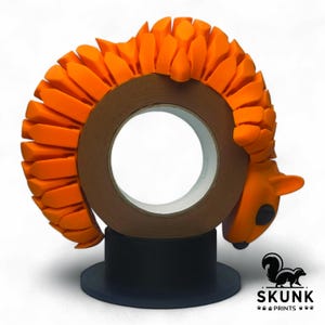 May include: A decorative tape dispenser featuring an orange squirrel design. The squirrel wraps around a roll of brown tape, mounted on a black base. The dispenser has a playful and unique aesthetic, perfect for a desk or office setting.