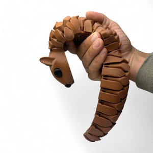 May include: A brown articulated deer toy with a segmented body and a deer head with black eyes and a black nose. The toy is held in a hand against a white background. The toy is made of plastic.